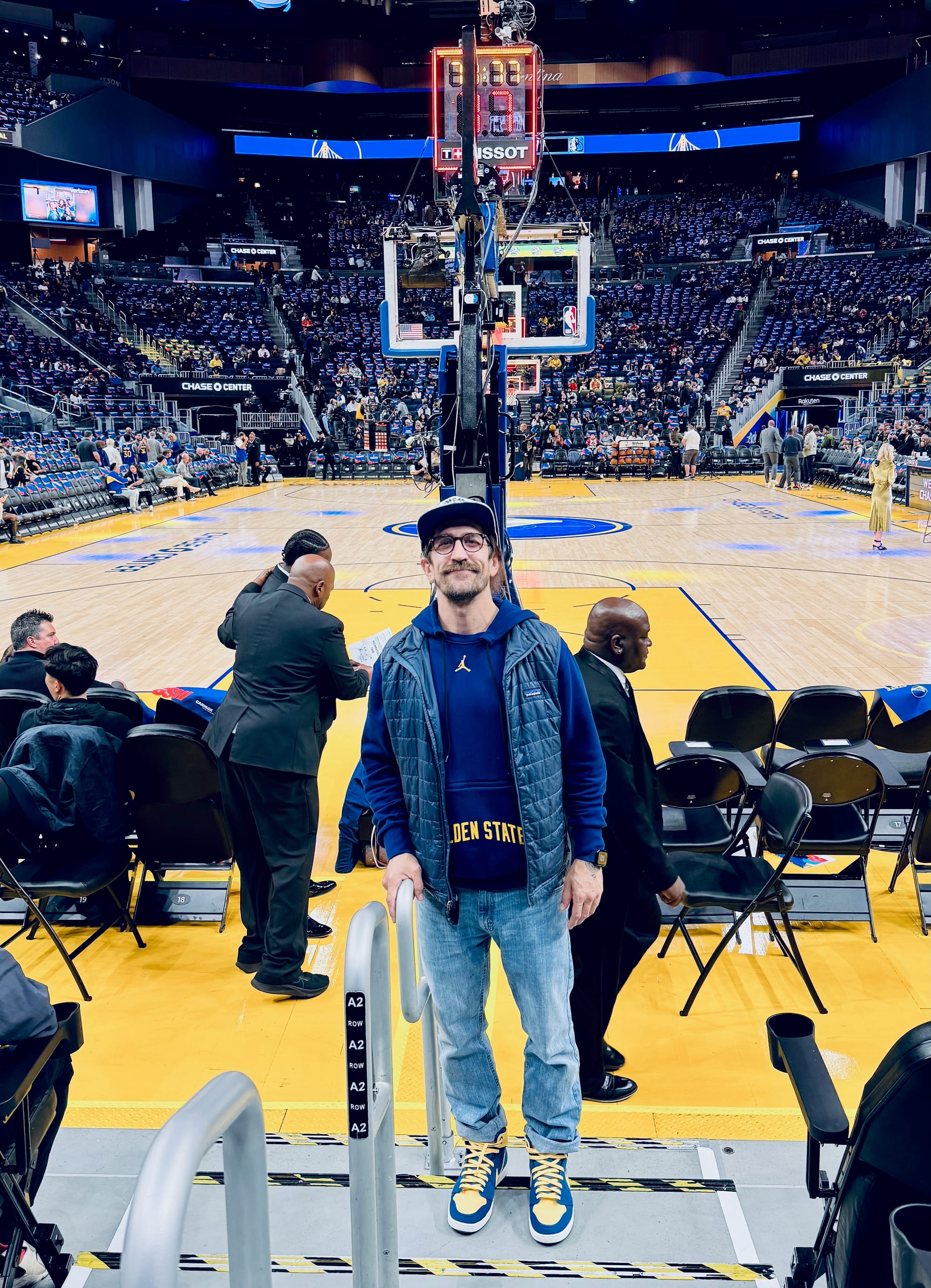 Tim standing courtside at Chase Center, home of the Golden State Warriors, wearing a Golden State hoodie, navy cap, and blue and yellow Air Jordan 1s that match the team colors. The hardwood court, basket, and Tissot shot clock are visible behind him, with the arena filling up in the background.